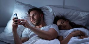 A man discreetly using his phone in the dimly lit bedroom while his unsuspecting wife is sound asleep, symbolizing potential evidence of an affair.